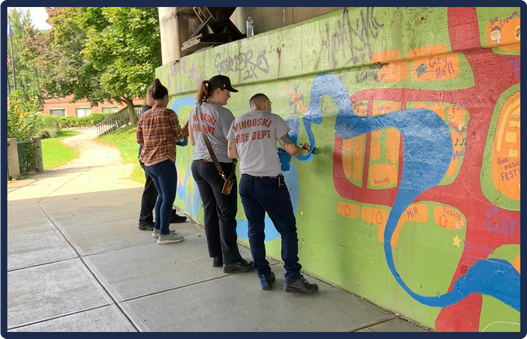 Three people painting on a wall under a bridge. The wall is covered in colorful paint, forming an abstract pattern. One person wears a white t-shirt with text on the back, while the others are in dark clothing. They use rollers to apply paint. The green surroundings include trees and grass