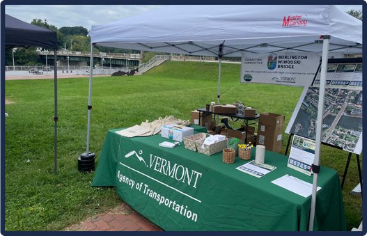 An outdoor information booth set up by the Vermont Agency of Transportation. The booth, covered by a white canopy tent, prominently displays a green banner with ‘VERMONT Agency of Transportation.’ On the table, various items like papers, maps, and informational materials are neatly organized. Behind the table, another banner highlights details about the Burlington Winooski Bridge Project. In the background, bleachers suggest a sports field or public event area.