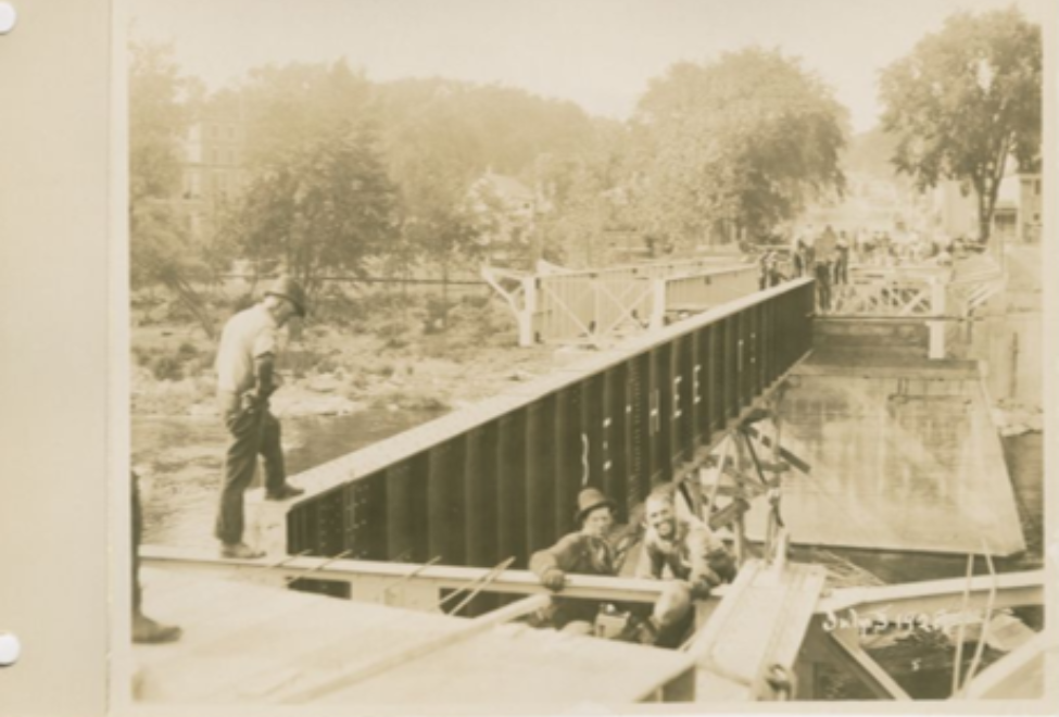 A historical photograph capturing individuals on and around a metal bridge over a river, surrounded by lush greenery.