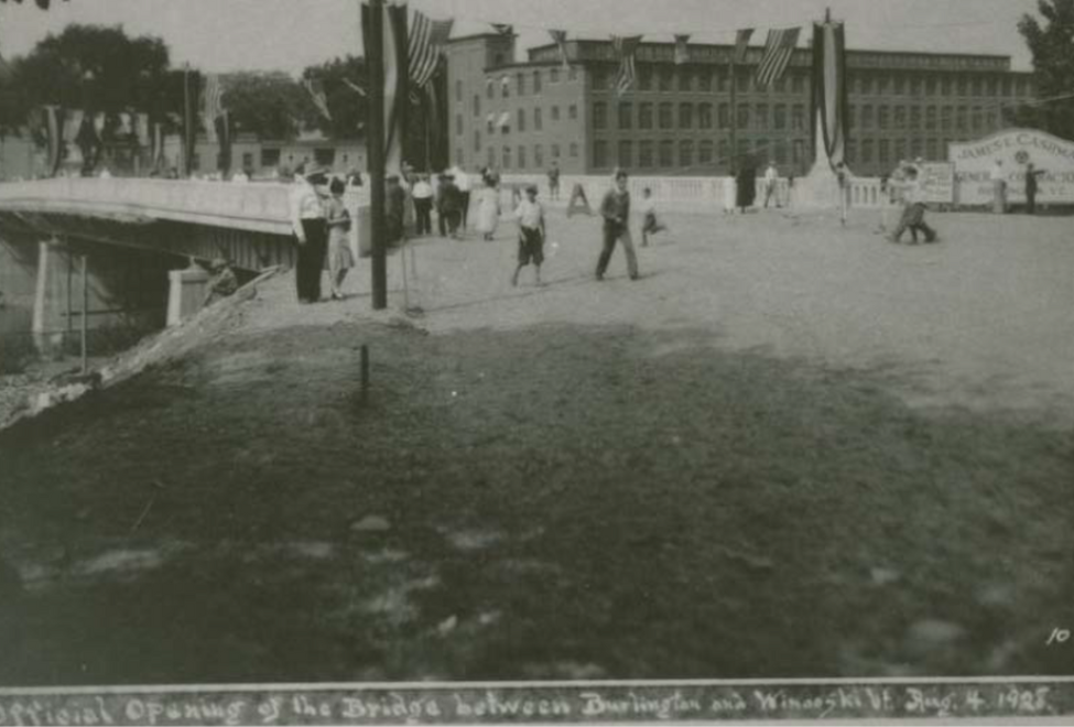 A historical black and white photo capturing the official opening of a bridge between Burlington and Winooski on August 3, 1928, with people gathered around and a large building in the background.