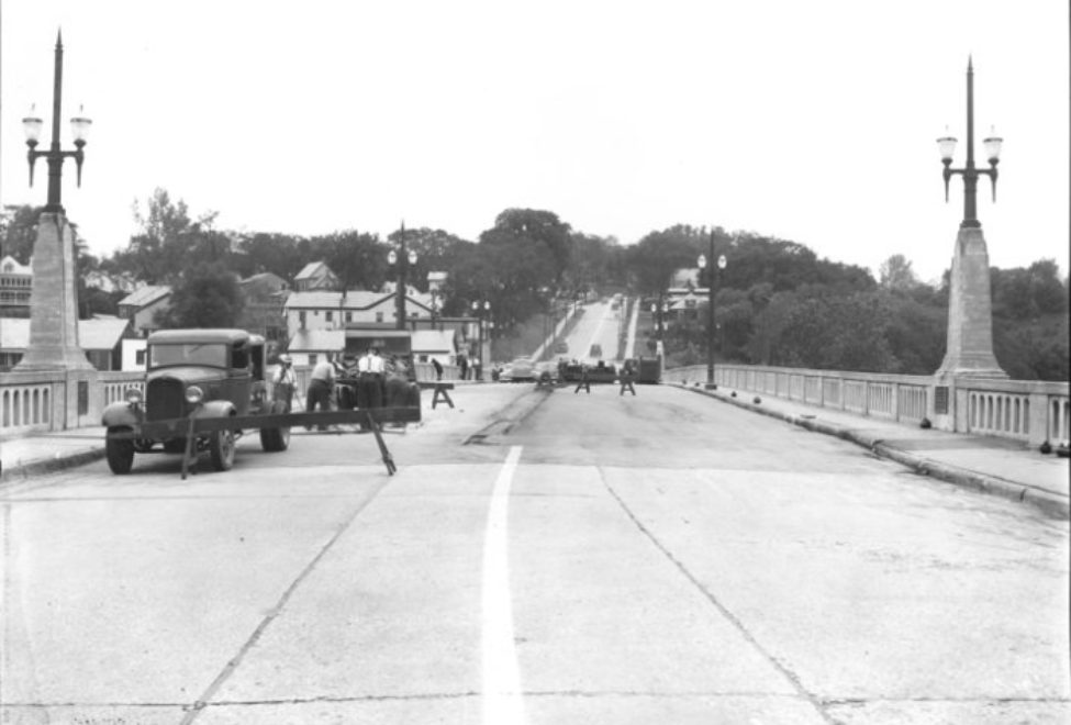 A historical photo of an old bridge with a vintage car, pedestrians, street lamps, and distant buildings. The bridge features ornate street lamps and concrete railings. A vintage car is parked on the left side of the bridge. Pedestrians are visible on the bridge. The road marking indicates two lanes. In the background, houses and trees on a hill create a suburban or small-town atmosphere.