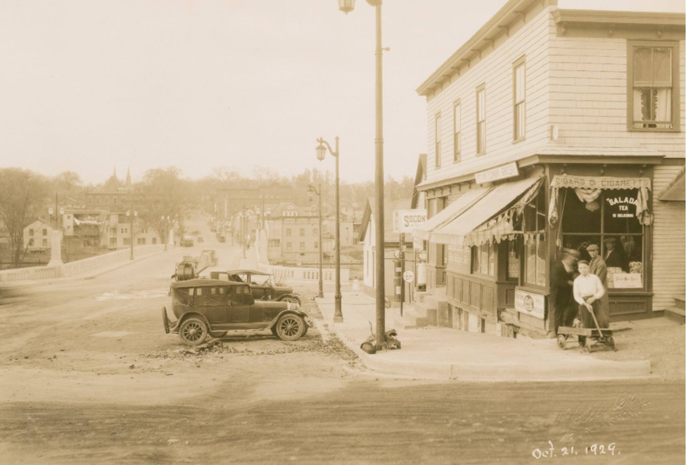 A historical photograph from October 21, 1949, showing a street scene. An old-fashioned car is parked on the left side of the image near a light pole. Two individuals stand in front of a store. The street appears to be under construction or repair, as it is not paved. Buildings line both sides of the street, which seems to be a mix of residential and commercial areas.